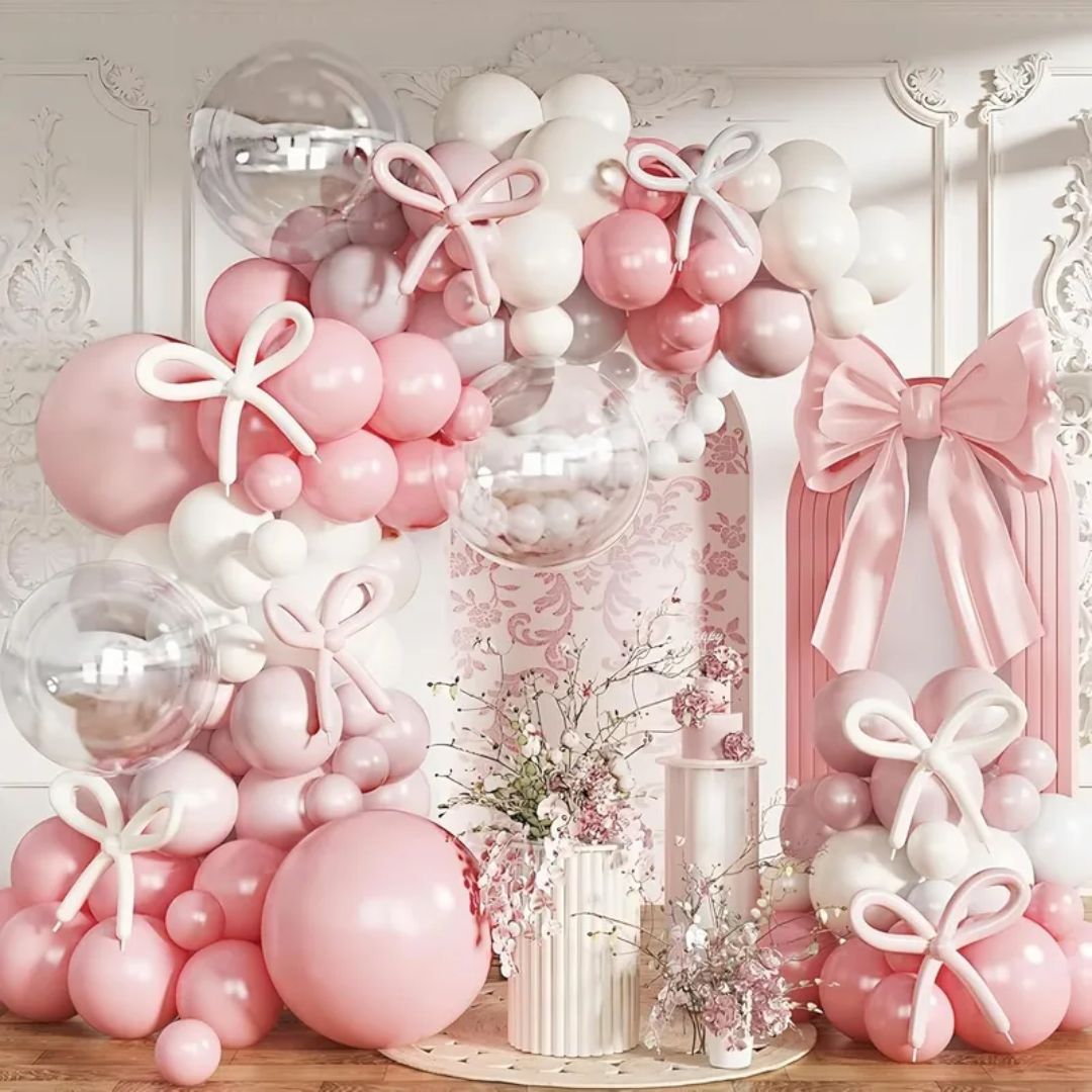 Pink and white balloon arch with decorative bows and flowers on a white wall.