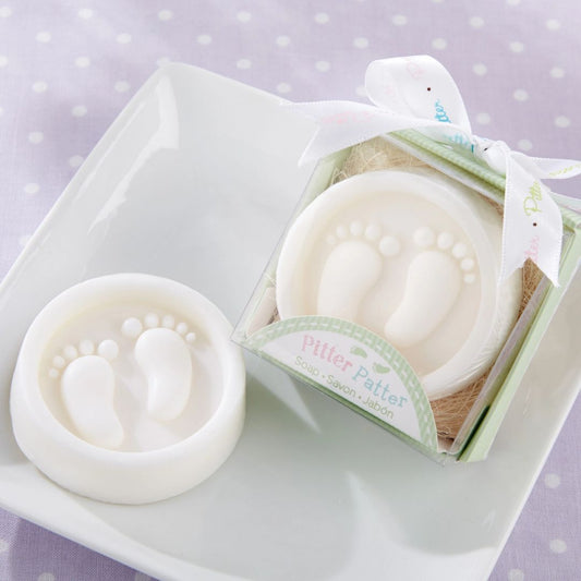 A white baby footprints soap and its matching white gift box with a ribbon, displayed on a white square plate over a light purple polka-dot fabric background. The box label reads 'Pitter Patter'.